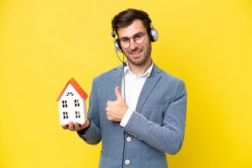 Young caucasian man holding a house isolated on white background giving a thumbs up gesture