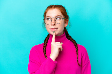 Young reddish woman isolated on blue background With glasses and looking up