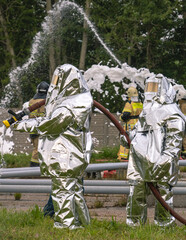 Firefighters in protective clothing use fire fighting equipment to supply water to extinguish the fire. In the background, firefighters extinguish a fire with foam. Firefighters extinguish the fire.