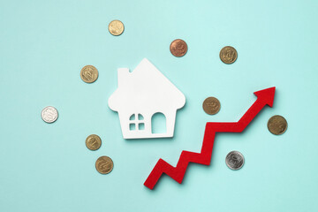 A white house model with a red upward arrow and scattered coins on a light blue background. Top...