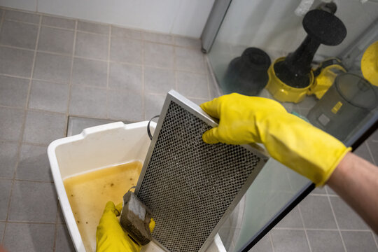 Person cleaning greasy metal range hood filter with sponge and rubber gloves over dirty water