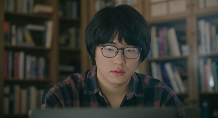 Student with glasses studying at laptop, books blurred background