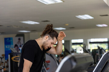 Young attractive fitness trainer feels exhausted and thirsty after too much exercise on a treadmill. A sport man gets dehydrated from treadmill running over long period of time in a fitness room