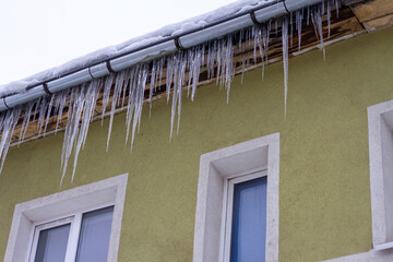 Long icicles hanging from roof gutter of residential building with green wall and white window frames.