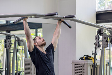 Young good looking physical trainer shows how to build upper part body by pulling up an equipment in a fitness room. A muscular sport man taking an exercise with fitness equipment to build his body.