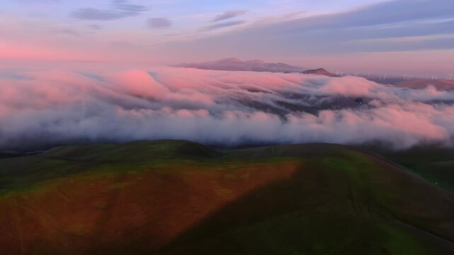 Aerial view of fog rolling over green hills at sunset in Altamont Pass, Livermore, California, USA. Mountains and wind turbines are visible in the distance.