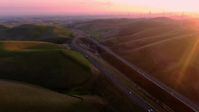 Aerial view of a highway cutting through rolling hills at sunset, with wind turbines in the background, showcasing renewable energy in Altamont Pass, Livermore, California, USA
