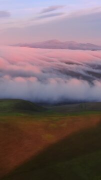 Aerial view of fog rolling over green hills at sunset in Altamont Pass, Livermore, California, USA. Mountains and wind turbines are visible in the distance.