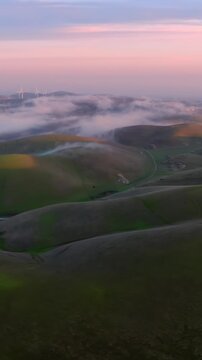 Aerial view of fog rolling over green hills at sunset in Altamont Pass, Livermore, California, USA. Mountains and wind turbines are visible in the distance.