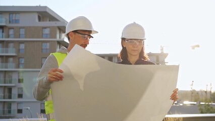 Two architects wearing safety hard hats are holding blueprint and discussing something on...