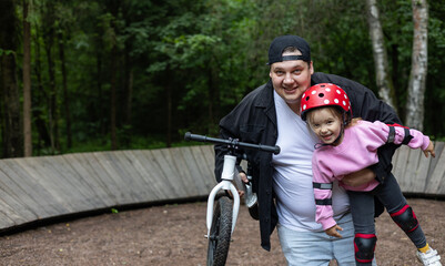 A father holds his smiling daughter in protective gear after riding a balance bike in the park,...