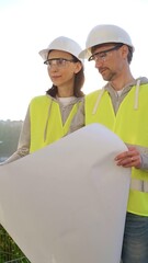 Two architects or engineers wearing safety hard hats and vests holding blueprint and discussing...