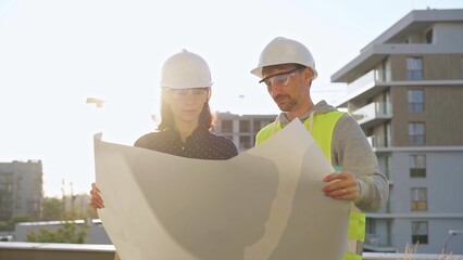 Two architects wearing safety hard hats are holding blueprint and discussing something on...