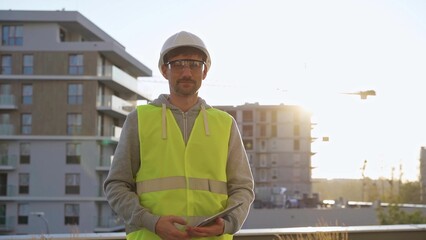 Construction site engineer holding tablet and wearing hard hat and safety vest with a sunrise at the background. Portrait video