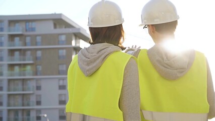 Man and woman architects or engineers wearing safety hard hats and vests holding blueprint and...