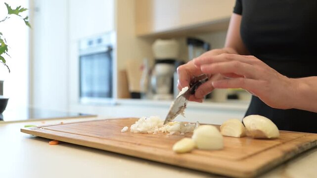 4K close-up of female hands slicing fresh onion and garlic on a wooden cutting board in a bright, modern home kitchen. Ideal for concepts of healthy home cooking with copy space