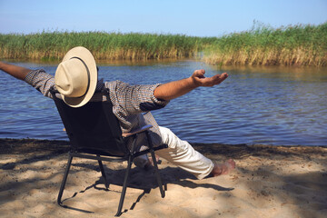 Man wearing hat enjoying freedom by the lake, relaxing on a folding chair with open arms