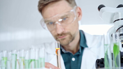 Man researcher with safety glasses is examining lab tube with a orange fluid in a modern...