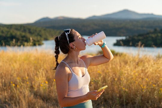 Young athlete drinking water and holding smartphone after training outdoors