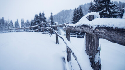 Rustic wooden fence covered in fresh snow in a peaceful winter mountain landscape, surrounded by pine trees and snowfall.