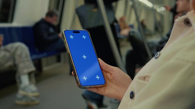A woman sits on a subway train, holding a smartphone with a blue screen. She is scrolling through content as the train moves through the urban environment.