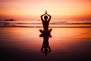 The silhouette of a woman in a seated yoga pose on the shore, reflected in the wet sand beneath a...