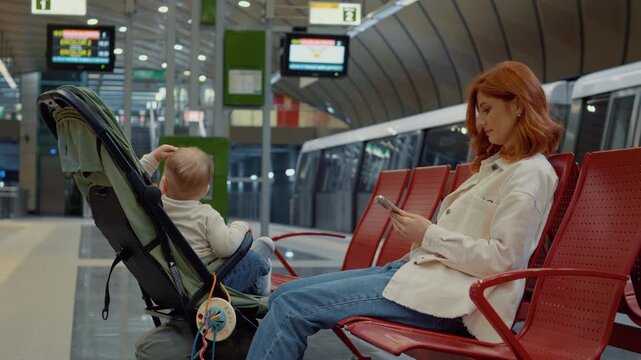 A young mother sits comfortably on a bench at the subway station, keeping her baby entertained while scrolling on her phone as they wait for the next train.