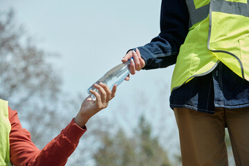 young adult handing plastic water bottle to another outdoors, both wearing reflective safety vests,...