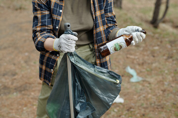 Young adult man wearing gloves collecting litter outdoors, holding empty glass bottle and placing...