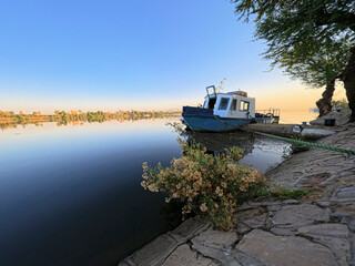 Small steel motor launch boat moored on the Nile river bank with rope mooring line and bush plant