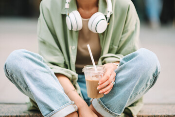 Casual dressed woman with headphones around her neck is enjoying her iced coffee in a plastic cup with straw while sitting outside