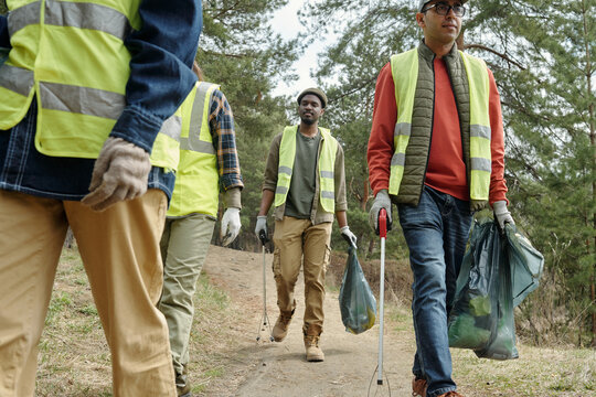 Group of diverse young adult men and women cleaning forest trail, collecting trash in plastic bags, wearing safety vests and gloves, participating in outdoor community cleanup event