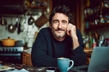 A middle-aged man with a beard sits thoughtfully at a kitchen table, surrounded by warm lighting, a laptop, and a cup of coffee.