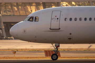 Obraz premium Primer plano del morro de un avión comercial con el piloto visible en cabina rodando por pista frente a la terminal del aeropuerto al atardecer