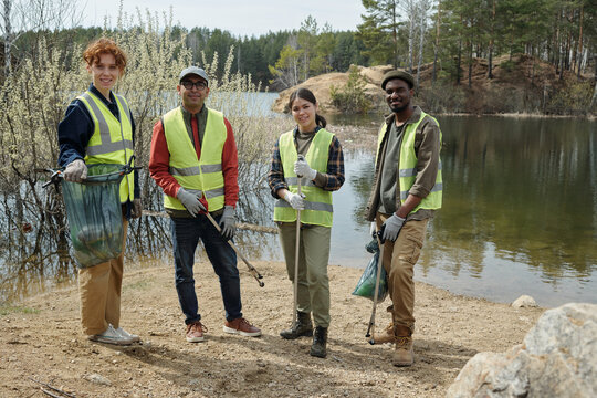 Group of young adult multiethnic men and woman standing outdoors near lake collecting trash, wearing reflective vests and gloves, participating in environmental cleanup activity