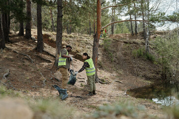 man and woman wearing safety vests collecting trash in forest near water, participating in...