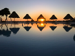 Sun umbrellas around an infinity swimming pool at luxury tropical hotel resort with orange dusk sunset and reflection