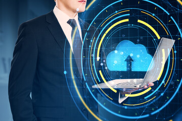 Businessman holding laptop with cloud upload icon and digital interface overlay on blue background, representing cloud computing and data transfer.
