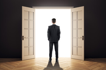 Businessman standing in dark room facing open door with bright light, symbolizing opportunity,...