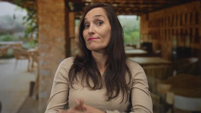 Woman with long hair and beige sweater gestures questioningly against a rustic restaurant terrace backdrop with wooden furniture and soft natural lighting outdoors