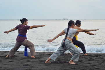 With the sea and a fishing boat behind, three teens stretch in a graceful yoga pose facing forward,...