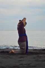 A teenage girl performs a graceful yoga stretch, kneeling on the beach with her right arm raised...