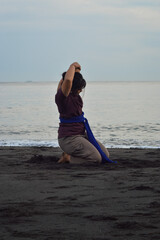 Facing the ocean, a teenage girl kneels in a yoga pose, left arm raised in motion, as waves roll in...