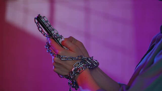 Shot showing a woman hands restrained by a metal chain with a mobile phone, symbolizing technological addiction.
