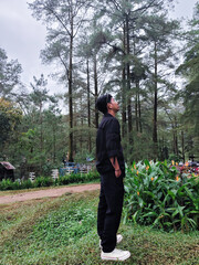 A young man in black clothes standing on grass and looking up, surrounded by pine trees and fresh greenery outdoors.
