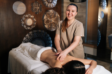Portrait of cheerful masseuse gently applying pressure to woman back during relaxing massage...