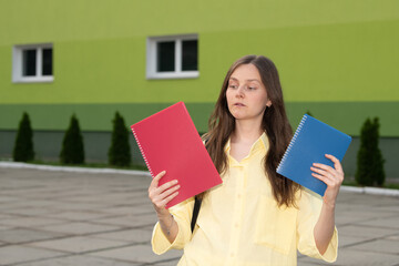 A young woman stands outdoors near a vibrant green building, thoughtfully holding a red notebook in...