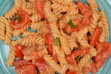 A close-up of a plate of fusilli pasta mixed with chunks of tuna, chopped tomatoes, and fresh basil