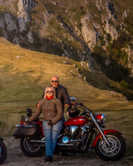 Smiling mature couple posing with a cruiser motorcycle in nature at sunset, enjoying a scenic...