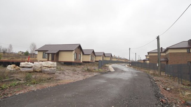 Rural road stretches between new houses rows standing on muddy slope. Asphalt leads uphill dividing plots and scattered construction bags